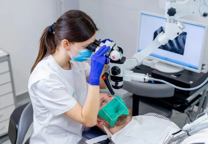 Dentystka w masce i rękawiczkach bada ząb pacjenta pod mikroskopem. Na ekranie widać zdjęcie rentgenowskie. To nowoczesne leczenie kanałowe, cena zabiegu zależy od skomplikowania.