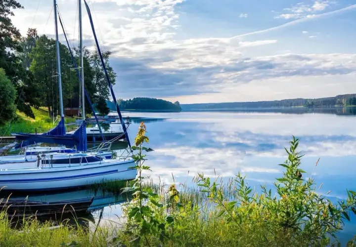 Żaglówki przy pomoście na spokojnym jeziorze. W tle las, a na wodzie odbijają się chmury. Mazury ciekawostki: idealne miejsce na relaks i żeglowanie.