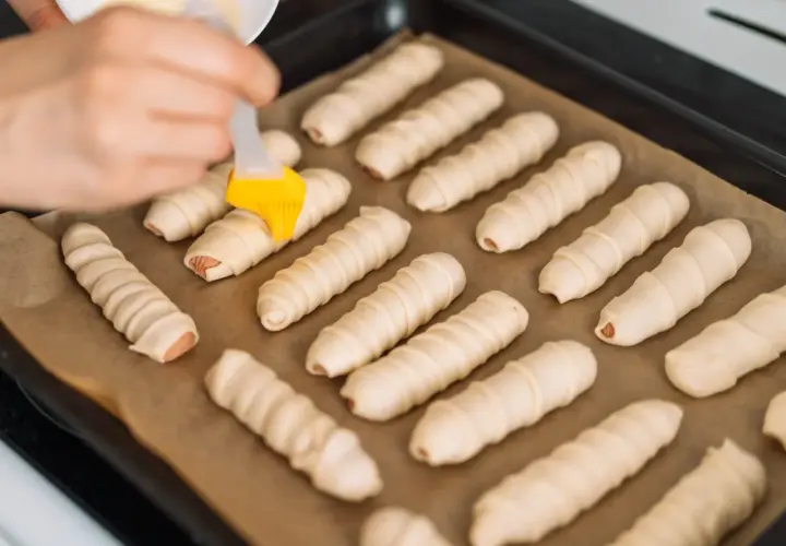 Ręka smaruje pędzelkiem parówki zawinięte w ciasto, przygotowując je do pieczenia. To prosty przepis na parowance.