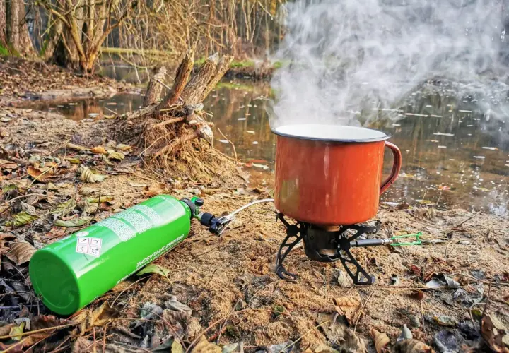 Kuchenka turystyczna z gazem i parującym kubkiem nad wodą. Różne rodzaje kuchenek pozwalają na gotowanie w plenerze.