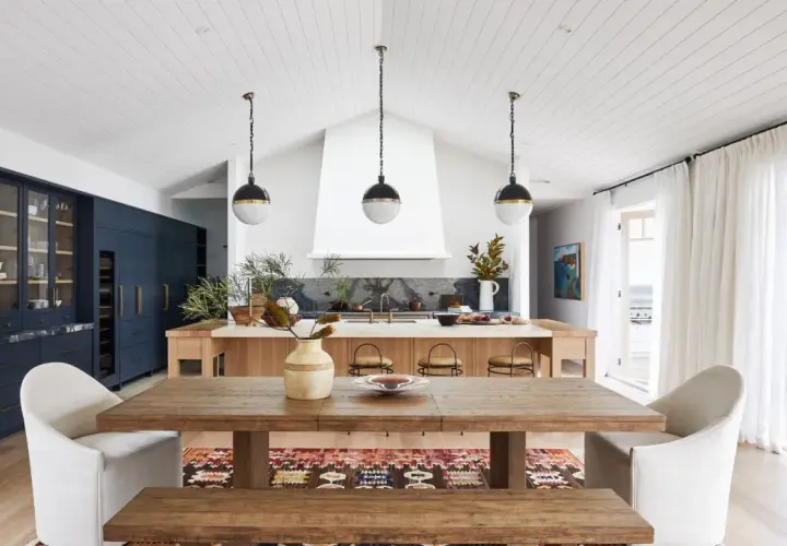 A modern lake house interior featuring a rustic wooden dining table, plush white chairs, and a sleek navy kitchen island.