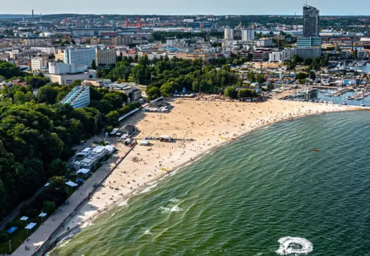 Słoneczny bulwar nadmorski Gdynia tętni życiem. Plaża pełna ludzi, a w oddali port z żaglówkami i miejska panorama.