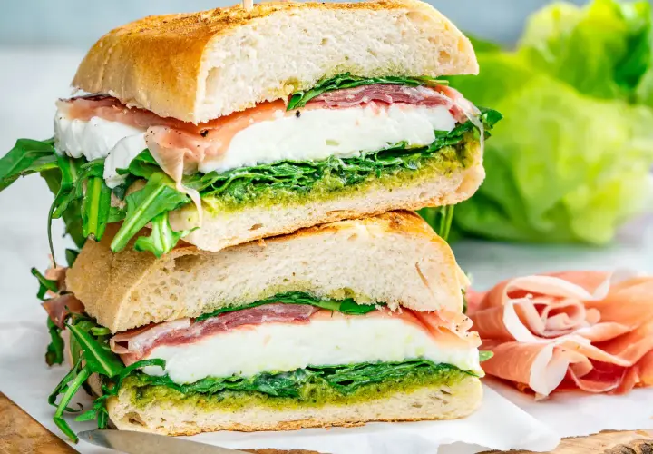 Dos bocadillos italianos apilados, rellenos de mozzarella fresca, prosciutto, rúcula y pesto, sobre pan rústico.