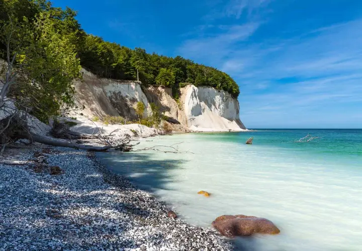 Weiße Kreidefelsen ragen steil auf, bewaldet von oben. Die Ostsee bei Sassnitz schimmert türkisblau, ein Kiesstrand lädt zum Verweilen ein.