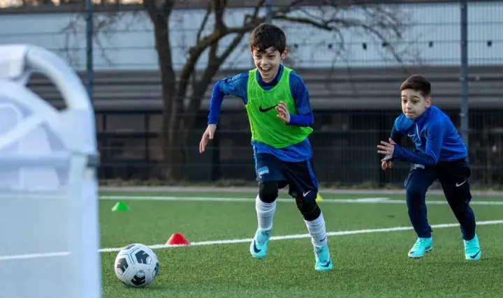 Zwei Jungen in Fußballtrikots trainieren auf einem Kunstrasenplatz. Sie sind voller Energie und Freude, während sie über das Feld rennen. Diese fussball spiele kinder sind ein toller Anblick!