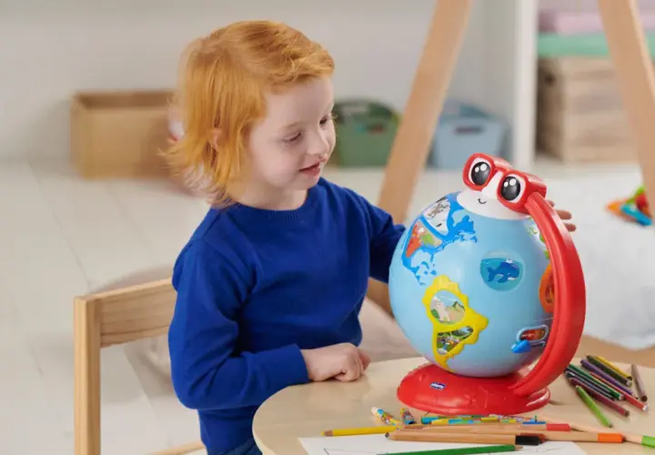 Un bambino di 5 anni con capelli rossi esplora il mondo con un globo interattivo, circondato da matite colorate.