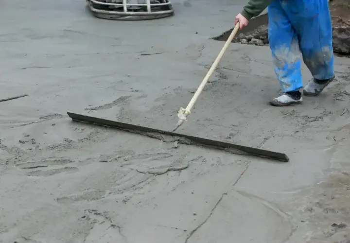 Pracownik w niebieskich ogrodniczkach wygładza świeży beton długą pacą. Trwa wygrzewanie posadzki.