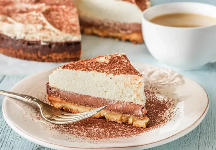 Pyszna tarta z mascarpone i czekoladą, posypana kakao, czeka na widelczyk. Obok filiżanka kawy.