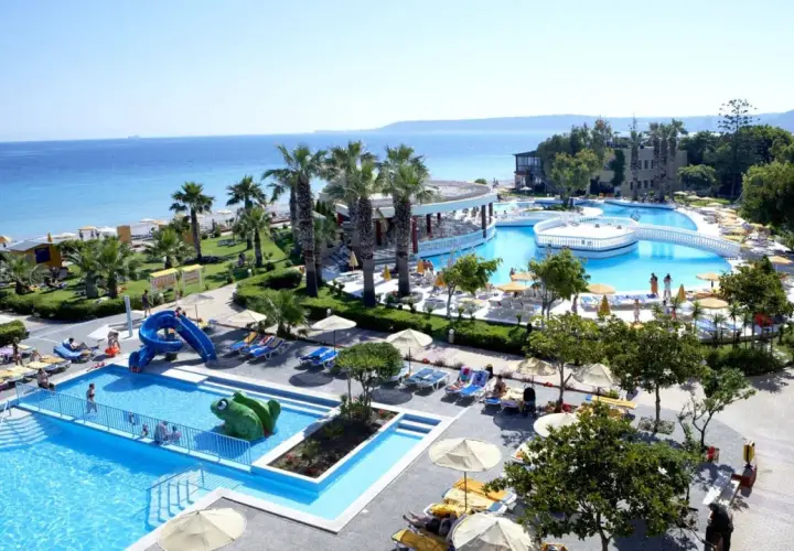 Słoneczna plaża i baseny w Ialyssos na Rodos. Dzieci zjeżdżają ze zjeżdżalni, dorośli odpoczywają na leżakach.