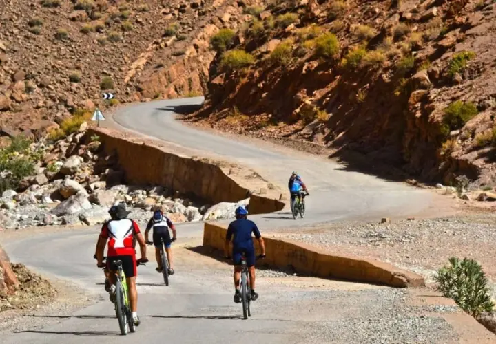 Ciclistas disfrutan de una aventura de **Marruecos en bici** por un camino sinuoso entre montañas rocosas y vegetación escasa.