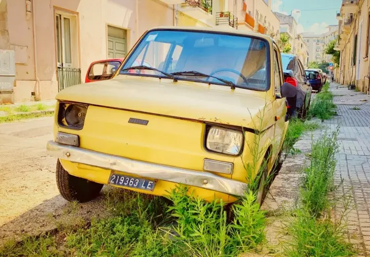 A vintage yellow Fiat, a symbol of driving in Puglia, sits parked on a sun-drenched street, overgrown with lush green grass.