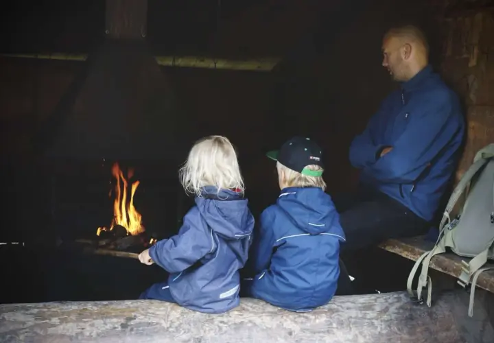 Två barn och en vuxen sitter vid en eld. En mysig paus under vandring med barn.