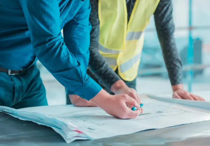 Deux hommes examinent des plans. L'un, en chemise bleue, pointe un détail avec un stylo. L'autre, en gilet jaune, pose ses mains sur les documents. Un constat huissier avant travaux est en cours.