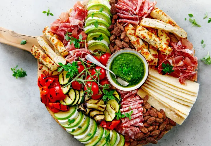 Tabla de antipasti italiani con prosciutto, salami, mozzarella, aguacate, tomates, pimientos y calabacín a la parrilla, acompañada de pesto y pan.