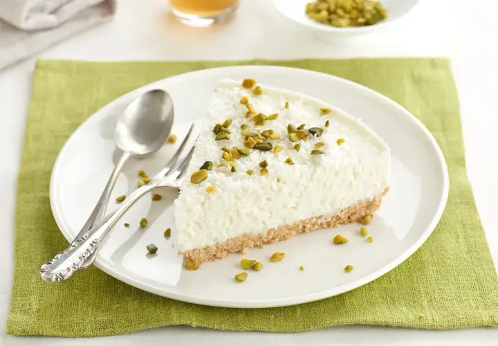 Rebanada de tarta de coco mousse, cubierta con coco rallado y pistachos picados, sobre plato blanco con cubiertos.