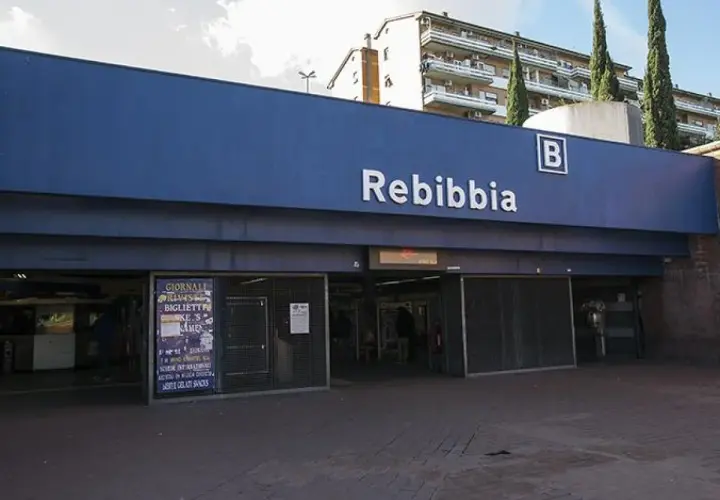 Ingresso della metro Rebibbia a Roma, con insegna blu e lettere bianche. Edifici residenziali sullo sfondo.