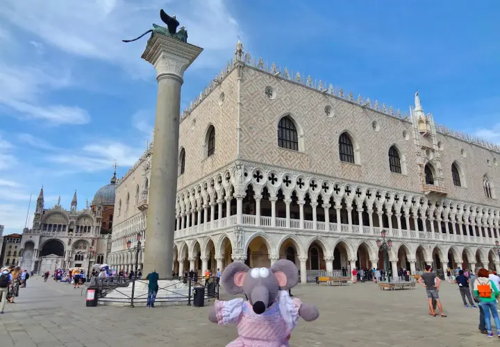 Une souris en robe rose devant l'entrée du Palais des Doges à Venise, avec la colonne de Saint-Marc et la basilique Saint-Marc en arrière-plan.
