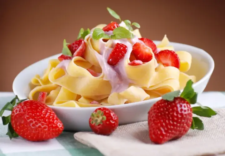 Pappardelle z kremowym sosem i kawałkami truskawek, ozdobione listkami mięty. Obok talerza leżą świeże truskawki.