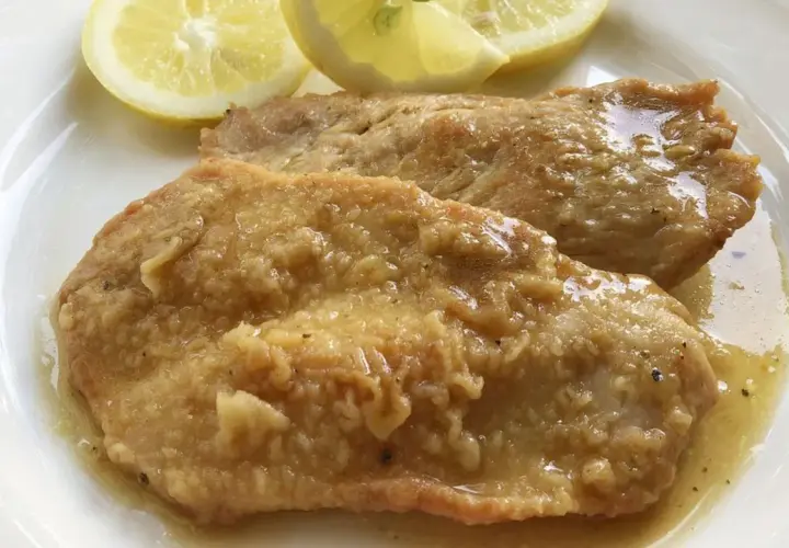 Deliziose scaloppine al limone, succulente e dorate, servite con spicchi di limone fresco su un piatto bianco.