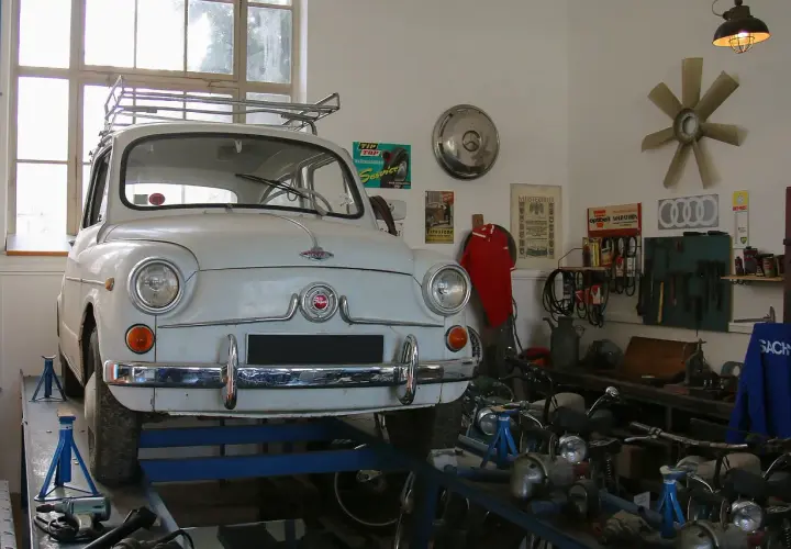 Biały Fiat 600 na podnośniku w warsztacie, gotowy do napraw. W tle narzędzia i motocykle, a nad nimi lampa zapewniająca oświetlenie kanału samochodowego.