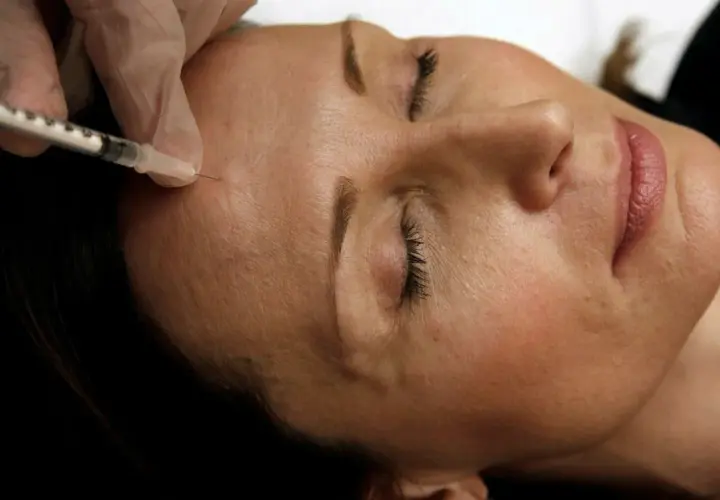 En hand i handske ger en injektion med en spruta i en kvinnas panna. Detta är en del av en behandling med botox på recept.
