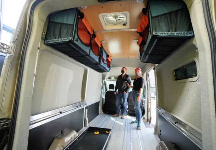 Aménagement fourgon par un professionnel : deux personnes admirent l'intérieur aménagé avec des rangements suspendus "Adventure Wagon" et un sol à rayures bleues.