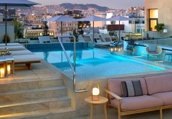 Piscina en la azotea del Barceló Raval con vistas a la ciudad al atardecer. Tumbonas, sofás y luces crean un ambiente acogedor.
