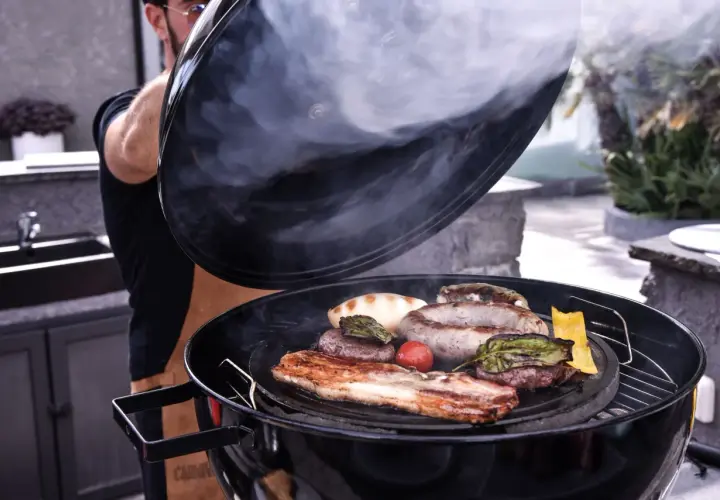 Un homme soulève le couvercle d'un barbecue fumant, révélant une sélection alléchante de viande au barbecue : saucisses, steaks, lard et légumes grillés.