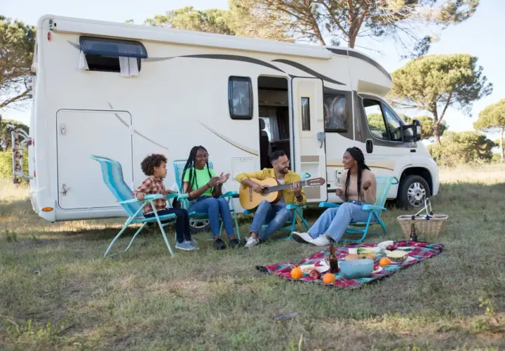 Familie genießt das Zelten mit Kindern. Ein Mann spielt Gitarre, während die anderen singen und lachen. Ein Picknickkorb und Obst liegen auf einer Decke.