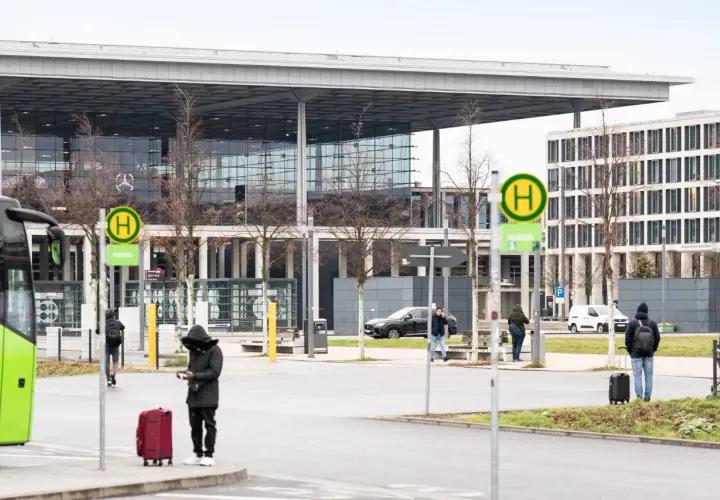 Przystanek autobusowy z zielonym autobusem, ludźmi z bagażami i nowoczesnym budynkiem w tle.