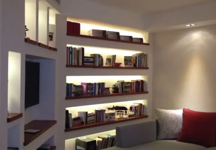 Libreria cartongesso angolare con illuminazione integrata, piena di libri e oggetti decorativi.