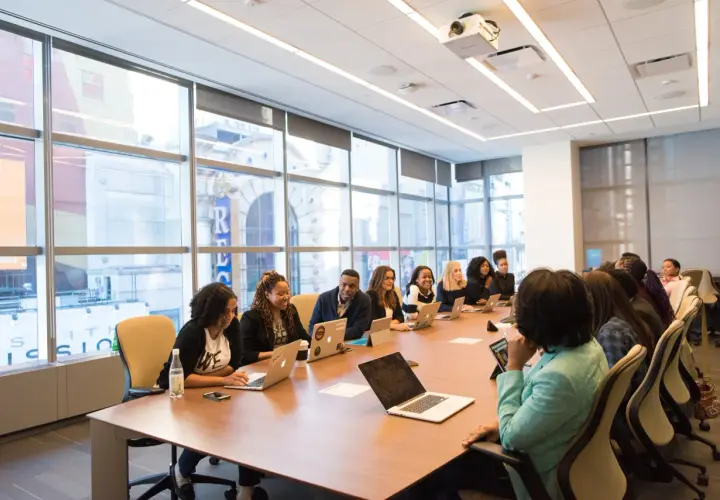 A diverse group of professionals engage in government leadership training, collaborating around a conference table with laptops and tablets.