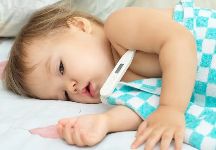 Bambino con termometro sotto l'ascella, avvolto in un asciugamano a quadri. La febbre che non scende bambini è una preoccupazione.