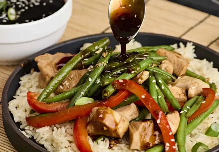 A spoon drizzles teriyaki sauce over a dish of rice, chicken, green beans, and red peppers, garnished with sesame seeds.