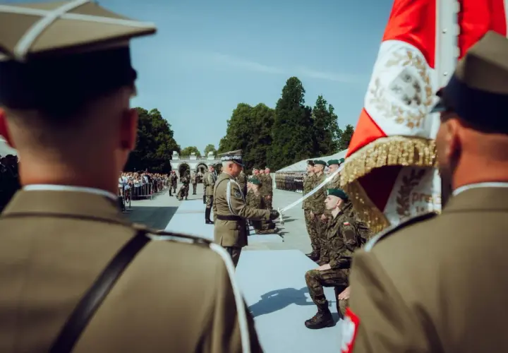Podczas uroczystości wojskowej, oficer wręcza szablę klęczącemu podporucznikowi. W tle stoją żołnierze i widzowie.