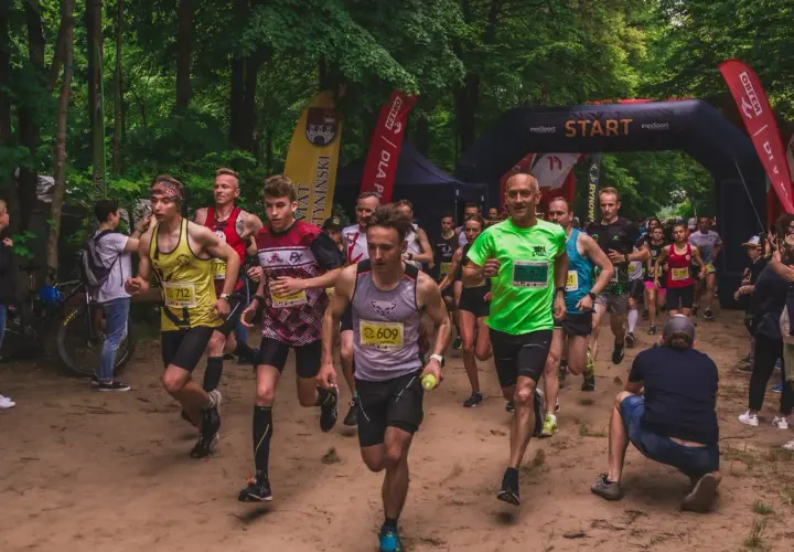 Uczestnicy biegu ultra trail startują w lesie. Widać łuk startowy, flagi i biegaczy w kolorowych koszulkach.