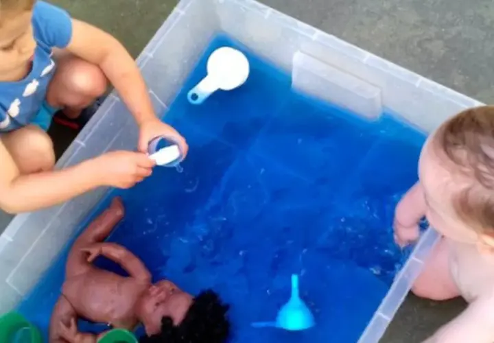 Niños de 3 a 4 años disfrutan de juegos acuáticos en el colegio, explorando con una muñeca y recipientes.