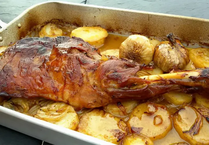 Paletilla de cordero al horno dorada y jugosa, acompañada de patatas asadas y ajos tiernos en una bandeja.