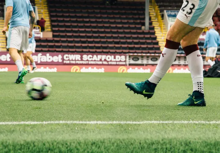 Piłkarz w białych spodenkach i zielonych butach kopie piłkę na boisku. W tle trybuny stadionu, charakterystyczne dla angielskich klubów piłkarskich.