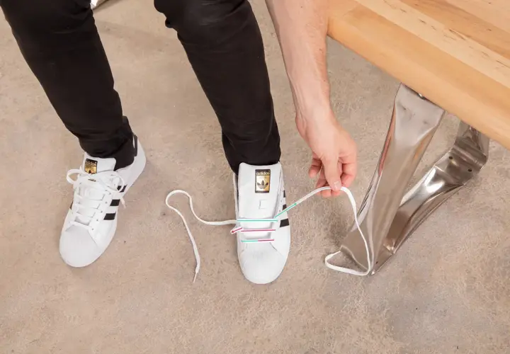 Instrukcja jak wiązać sneakersy: białe buty Adidas z czarnymi paskami, osoba zawiązuje sznurówki.