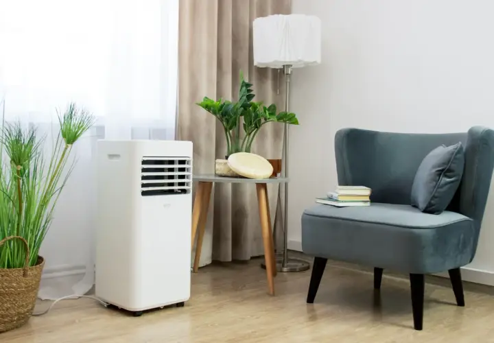 Biały klimatyzator przenośny stoi obok rośliny w wiklinowym koszu. Wygodny fotel z książkami i poduszką dopełnia wnętrze.