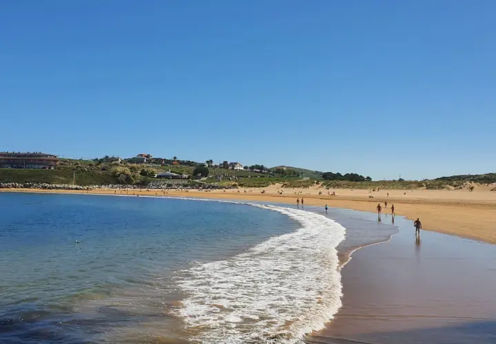 La **playa de Cuchía** se extiende bajo un cielo azul, con gente paseando por la arena dorada y el mar acariciando la costa.