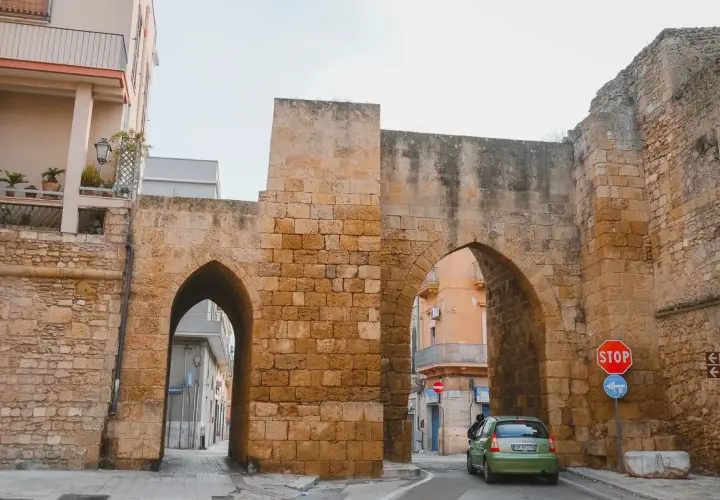 Antigua puerta de piedra con arcos, un coche verde pasa. Un lugar perfecto para un brindis por la historia.