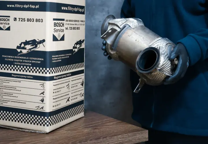 Osoba w rękawiczkach trzyma element układu wydechowego, prawdopodobnie filtr DPF. Obok stoi karton z logo Bosch Service, informujący o regeneracji filtrów DPF w dieslu.