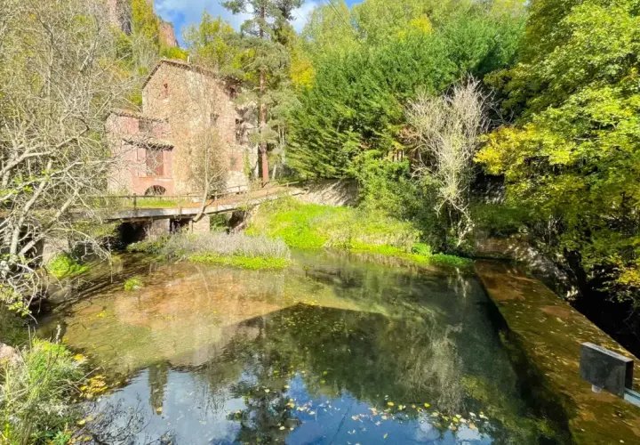 Antigua edificación de piedra junto a un río cristalino, rodeada de frondosa vegetación otoñal. Un puente de madera cruza el agua, conectando con las **fonts del Llobregat**.