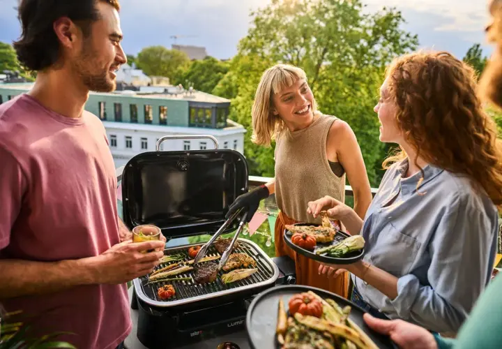 Vänner njuter av en grillfest på balkongen med en bärbar kolgrill. Saftiga biffar och grönsaker grillas medan solen går ner.