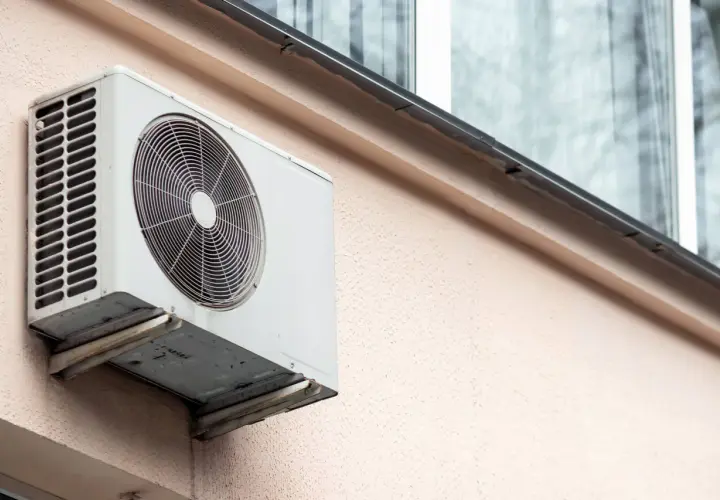 Klimatyzacja w bloku, biała jednostka zewnętrzna zamontowana na jasnoróżowej ścianie, z widocznym wentylatorem i kratką.