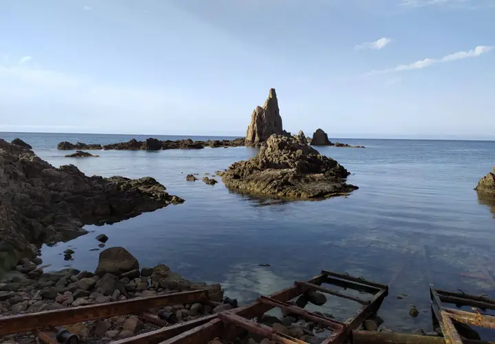 Rocas escarpadas emergen del mar azul, evocando el misterio del arrecife de las sirenas. El agua cristalina revela el lecho rocoso y restos oxidados de estructuras antiguas.