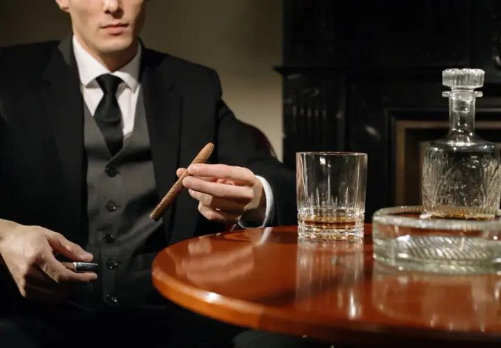 Mężczyzna w garniturze trzyma cygaro, obok szklanka whisky i karafka. Dobór whisky do cygar: intensywność i profil aromatu.