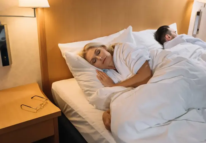 Frau schläft friedlich im Bett, nach dem Mittagsschlaf benommen. Neben ihr ruht ein Mann. Brille liegt auf dem Nachttisch.
